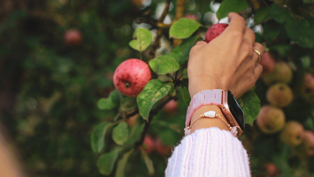 Weibliche Hand mit einem rosa Apple Watch Armband greift nach einem roten Apfel, der am Baum hängt.